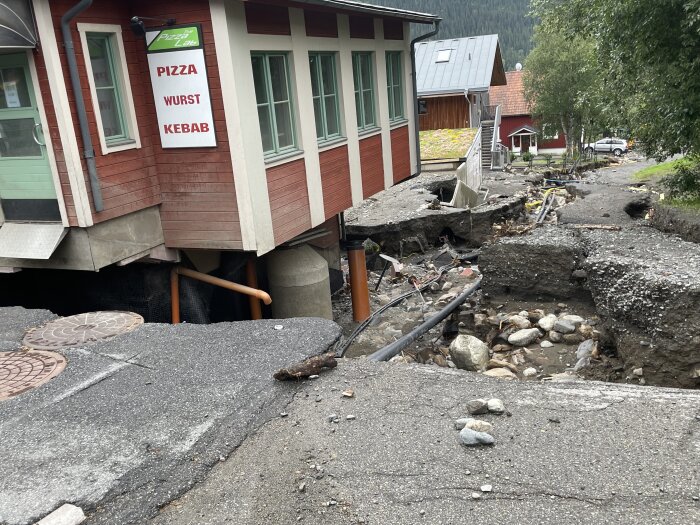 Erosion eller jordskred skadat byggnad och väg, synliga rör och stenar, restaurangskylt med "PIZZA WURST KEBAB".