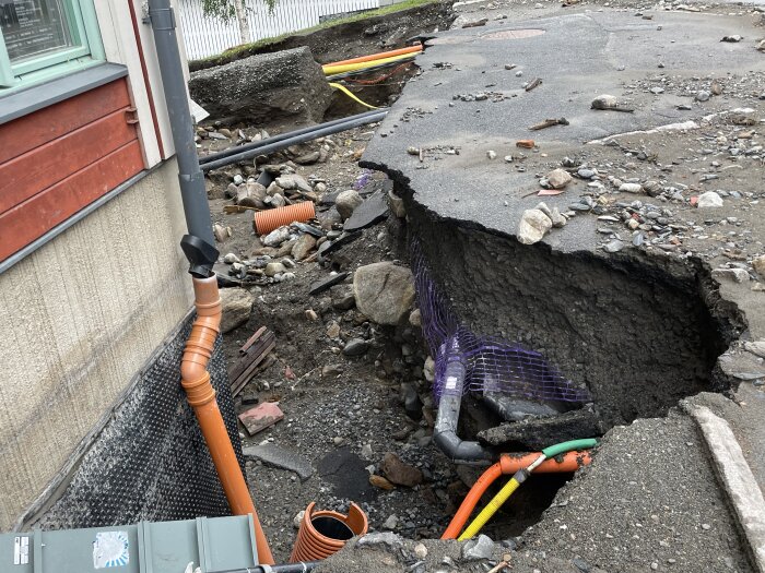 Asfalterad yta kollapsat och bildat en stor grop vid husgrund, exponerar rör och infrastruktur.