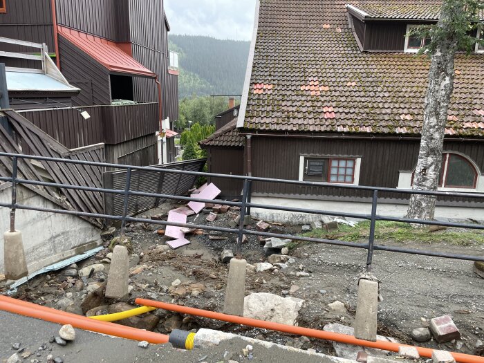 Byggarbetsplats med förstört staket, isoleringsmaterial på marken, byggnader i bakgrunden, staket i förgrunden, rör på marken.