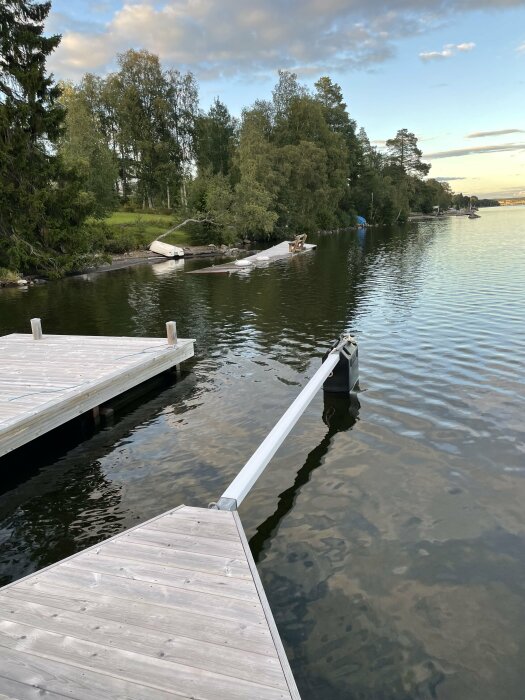 Träbrygga sträcker sig över lugnt vatten, träd, båtar och skymning. Ser ut som en fridfull kväll vid sjön.