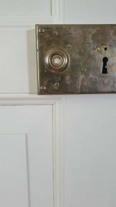 Vit dörr med metallisk låsbleck, låskista, vred och nyckelhål, slitet utseende, ingen synlig dörrhandtag.