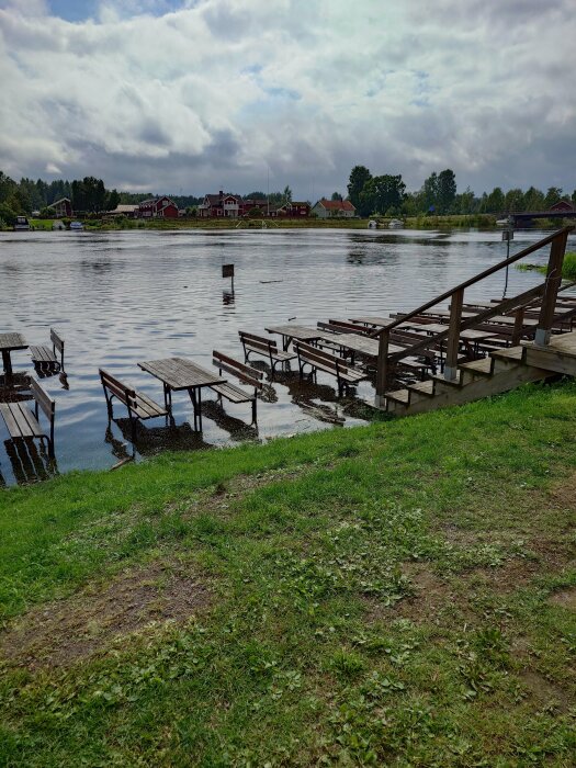 Sjöutsikt med bryggor, molnig himmel, grön gräsmatta, traditionella röda trähus i bakgrunden.