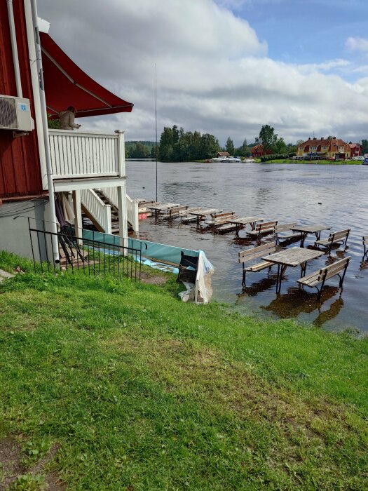 Röd stuga med veranda vid sjö, molnig himmel, bryggor, båt, grönt gräs, lugnt vatten, småstadsbebyggelse i bakgrunden.