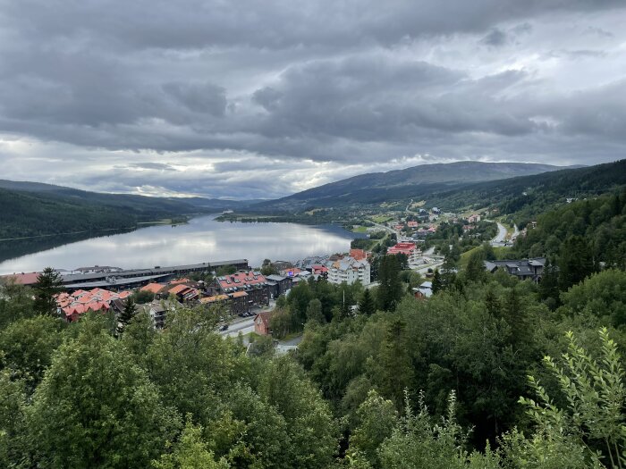 En småstad vid en sjö omgiven av skog och berg under en molnig himmel.
