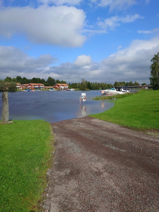 Översvämmad väg och gräsplätt, sjö, båtar, röda hus, molnig himmel, lugnt.