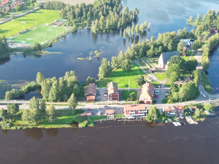 Flygfoto över flod, bebyggelse, grönområden, idrottsplan och båtar. Soligt och grönt landskap med reflektioner i vattnet.