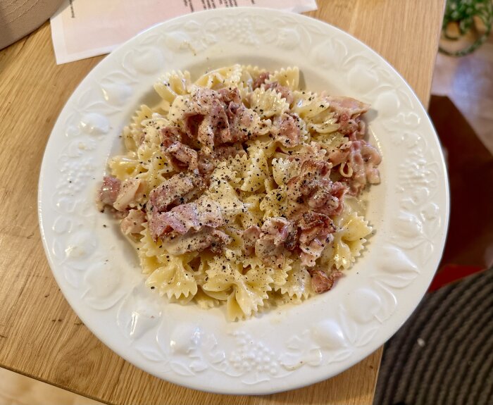 En tallrik med pasta och bacon toppad med svartpeppar på ett träbord.