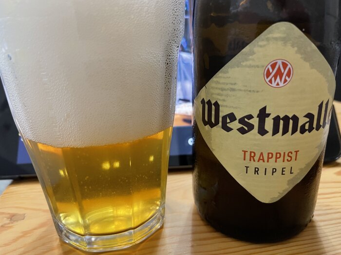 Glas med öl, tjockt skum, flaskan Westmalle Trappist Tripel, underlag av trä.