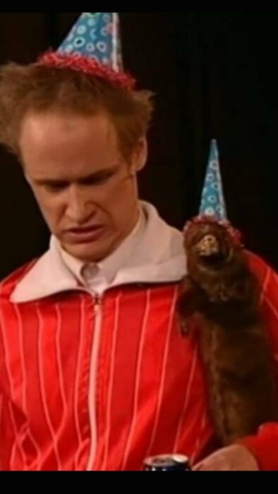 Man i röd jacka, festhatt, med ledsen min. Otter har också hatt. Feststämning med humoristisk twist.