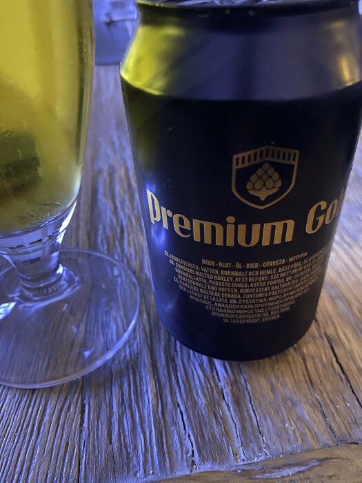 Ölburk med "Premium Gold"-text, glas med gul dryck, träbord, närbild, mörkt rum.