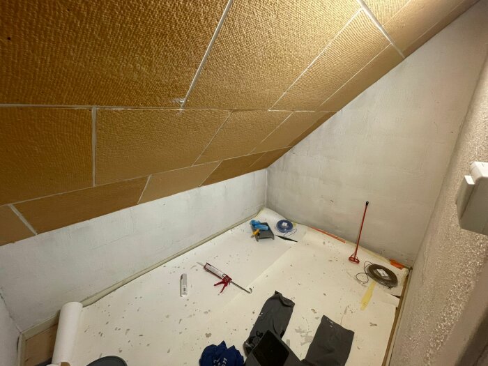 Ett oinrett hörnrum med lutande tak, byggmaterial på golvet, vit vägg, under renovering eller konstruktion.