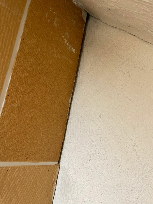 Hörnet av ett rum med beige vägg, vitmålad karm och brunt golv.