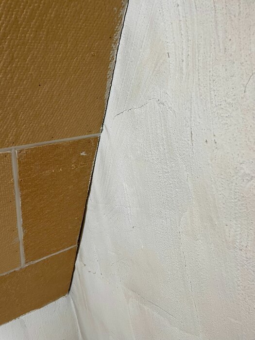 En vägg med beigea kakelplattor möter en vitmålad yta med sprickor vid skarven.
