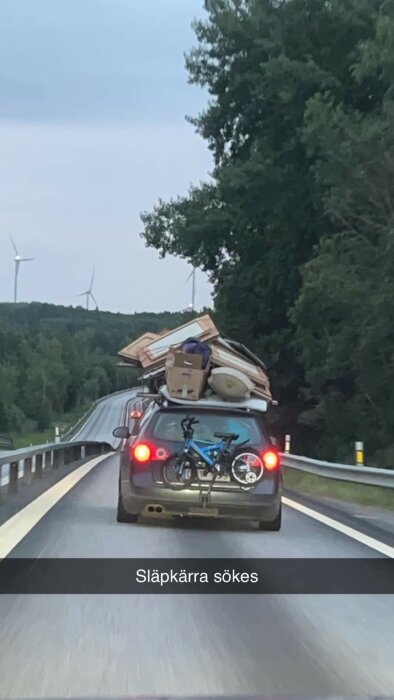 Bil överlastad med möbler, cykel bakpå, kör på väg, vindsnurror i bakgrunden, text "Släpkärra sökes" humoristisk.