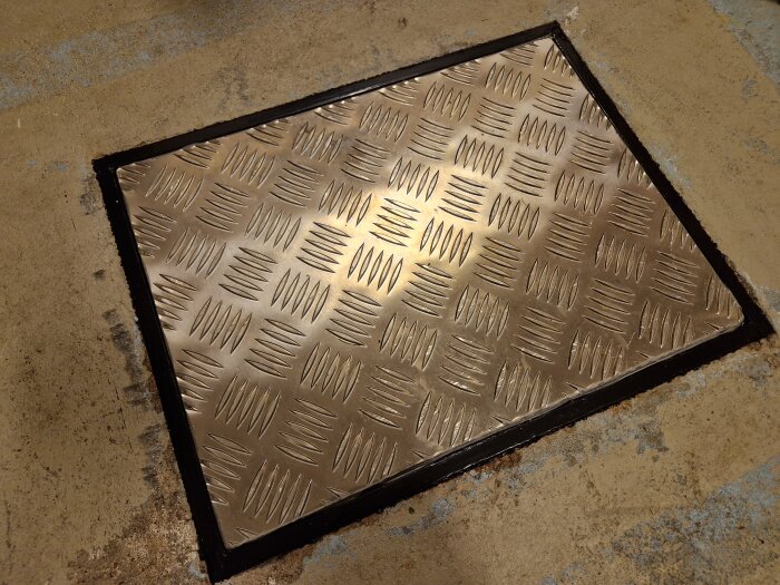 Metallgaller med diamantmönster i betonggolv, nedslitet, används för ventilation eller tillträde.