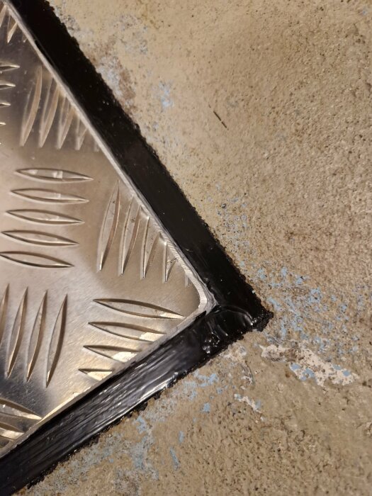 Metallkant med mönster mot betonggolv, svart tätningsmassa, slitna ytor, närbild.