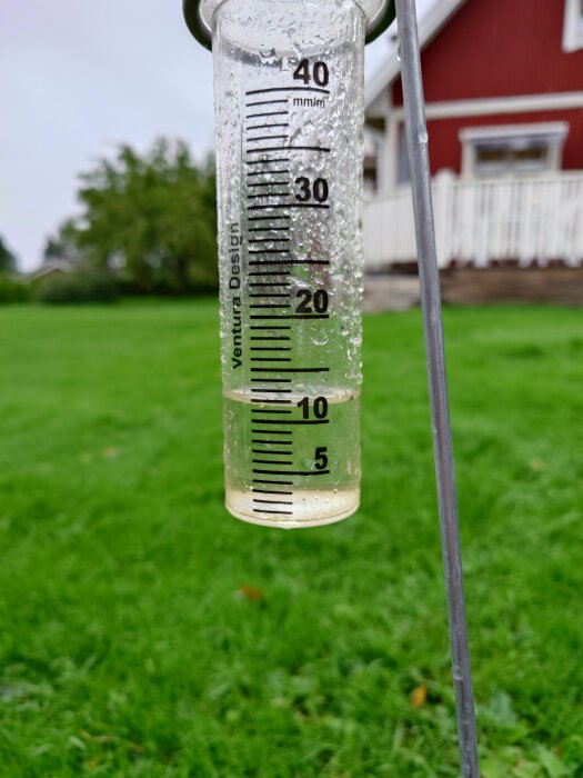 Regnmätare på gräsmatta visar nederbörd, hus och träd i bakgrunden. Cirka 5 mm regn.