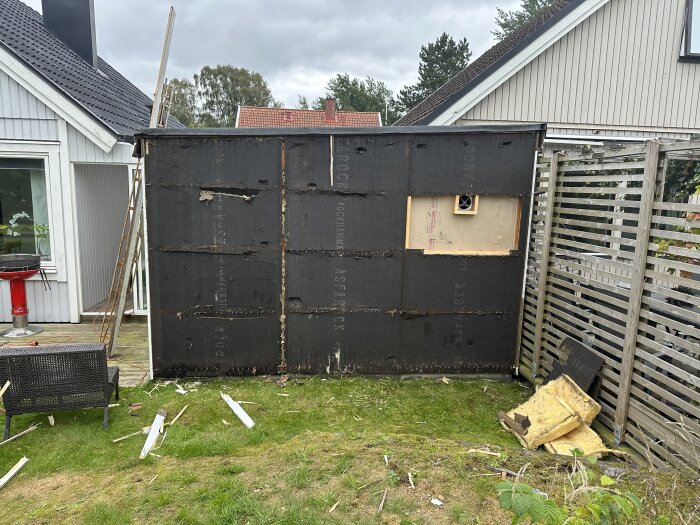 En förfallen skjul med svarta väggar, reparation pågår, bråte på gräsmattan, bostadshus i bakgrunden.