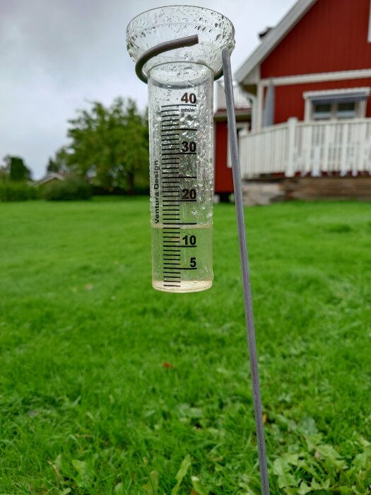 Regnmätare i trädgård med ungefär 5 mm nederbörd, röd stuga i bakgrunden, gräsmatta, dagtid.