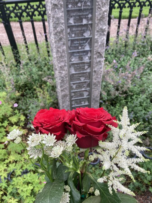 Gravsten med namn, röda rosor, vita blommor, grönska, svart stängsel i bakgrunden.