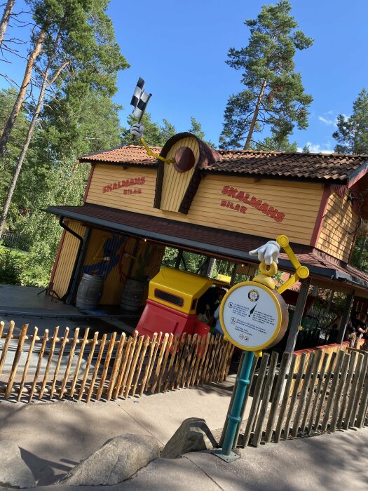 Temaparkskiosk betitlad "Skalmans Bil & Båt", trästaket, träd i bakgrunden, stolpe med skylt, lekfull arkitektur.