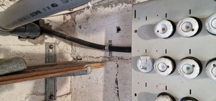 Elrör, kablar, säkringsskåp och trälister på en ojämn betongvägg. Oorganiserade installationer, slitage, åldrande infrastruktur.