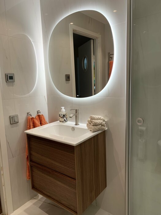 Modernt badrum med upplyst spegel, träkommod, handfat, och orange handdukar.