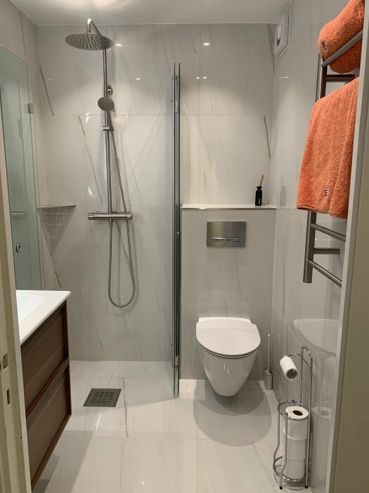 Modernt badrum med duschkabin, toalett, vita kakelväggar och terrakottafärgade handdukar.