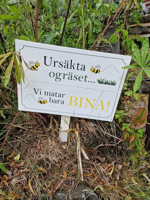 Skylt bland vegetation: "Ursäkta ogräset... Vi matar bara BIN!", med bin-illustrationer, naturbakgrund.