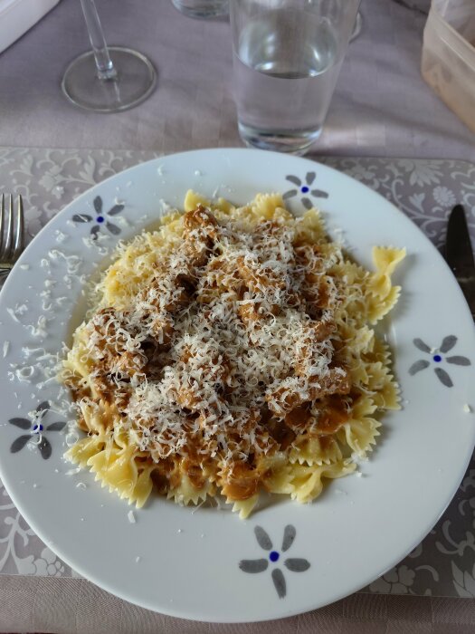 Farfalle-pasta med köttsås, toppad med riven ost, på blommönstrad tallrik, med glas vatten i bakgrunden.