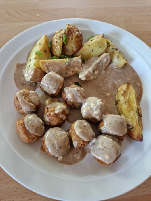 En tallrik med köttbullar, gräddsås och klyftpotatis, garnerad med örter.