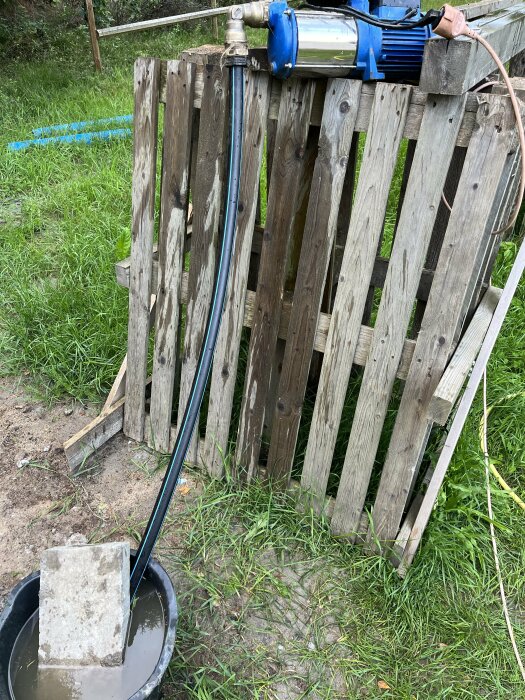 En vattenpump monterad på en gammal trästaket med slangen i en betongblock-fylld hink, omgiven av gräs.