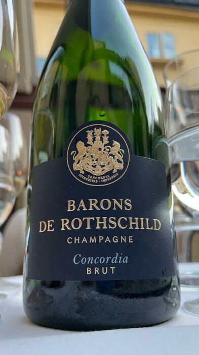 En flaska champagne med etikett "Barons De Rothschild Champagne Concordia Brut", bakgrunden visar ett bordsarrangemang.