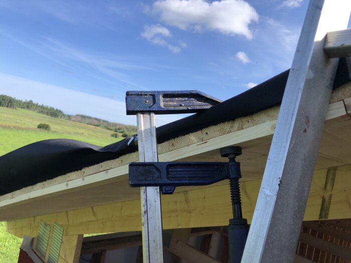 Två skruvstäd pressar trädelar under konstruktion utomhus med grön mark och blå himmel.