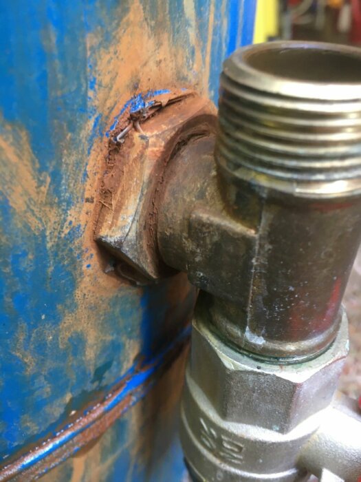 Metallrör med gänga fäst på blå behållare, rost, blå färgflagor, nötningsmärken, möjlig tätningskomponent synlig.