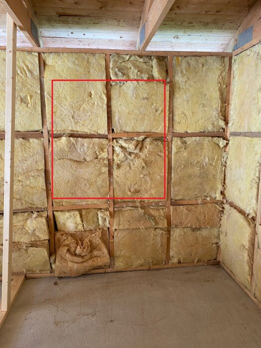 En vägg med fiberglasisolering. Röd ram indikerar område. Ojämn isolering nedtill. Oavslutad konstruktion. Träreglar synliga.