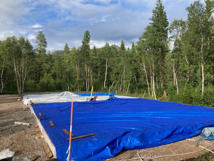 Byggarbetsplats, blå presenning, trästöttor, skogsbakgrund, molnig himmel, outvecklat landskap, grus, spridda byggmaterial.