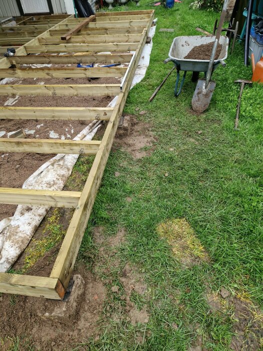 Träramkonstruktion för däck eller altanbygge på gräsmatta med skottkärra och verktyg.
