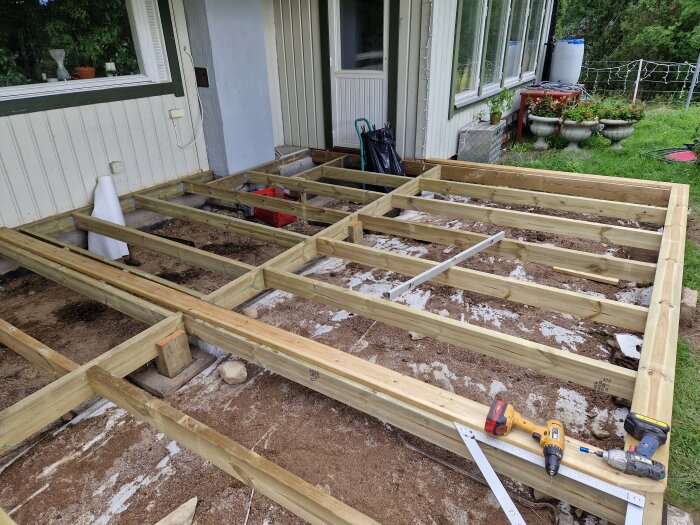Grundläggning träramar för däckbyggnation nära hus, verktyg och byggmaterial synliga, pågående arbete.