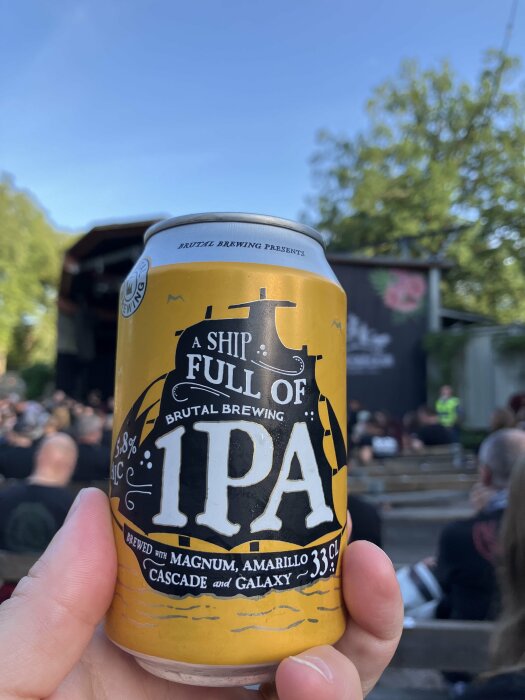 En burk IPA-öl hålls upp framför en folksamling vid en utomhuskonsert eller evenemang på dagen.