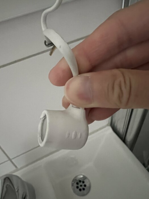 Hand håller brutet duschhandtag över handfat; skadad badrumsutrustning, vit kakelbakgrund.