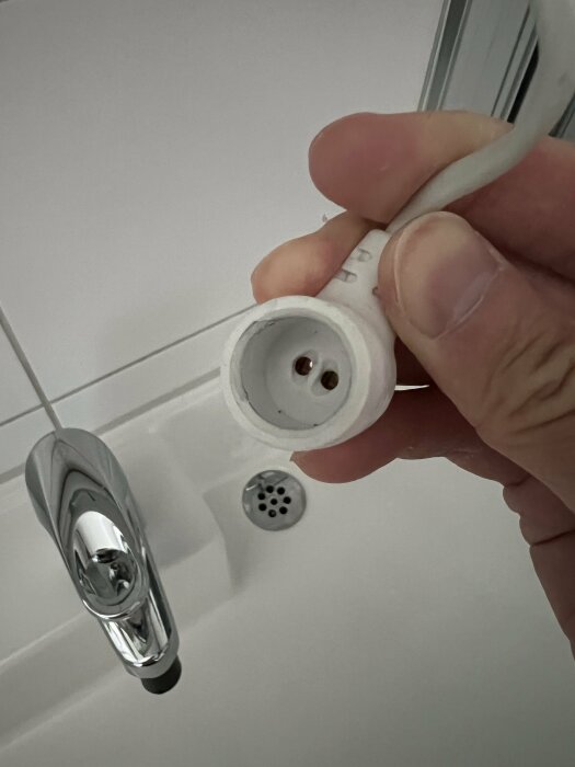 Hand håller en vit elkontakt nära en kran och en avloppsventil på ett badrum.