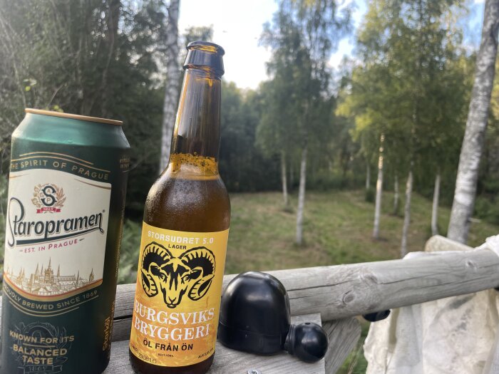 Ölburk och flaska på trädäck med skog i bakgrunden. Hörlurar och tyg på räcket. Utomhus, avslappnad atmosfär.