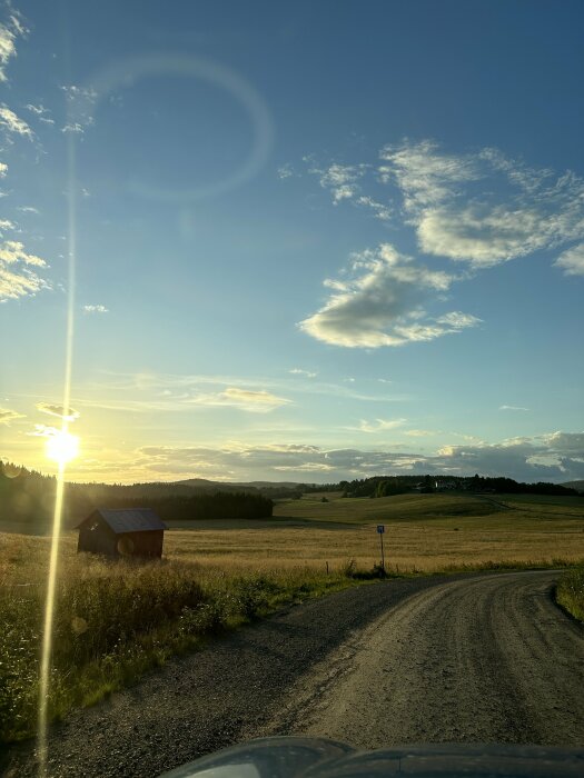 Lantväg vid solnedgång, grusväg, gyllene fält, röd lada, blå himmel med moln, solstrålar, naturlig skönhet.