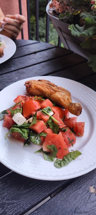 Grillat kycklinglår och vattenmelonsallad på tallrik utomhus. Modernt, fräscht, somrigt. Oskarp person i bakgrunden.