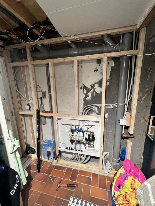 Ett ofärdigt utrymme med synlig rördragning, elcentral, och isolering. Verktyg och byggmaterial syns. Pågående renovering.