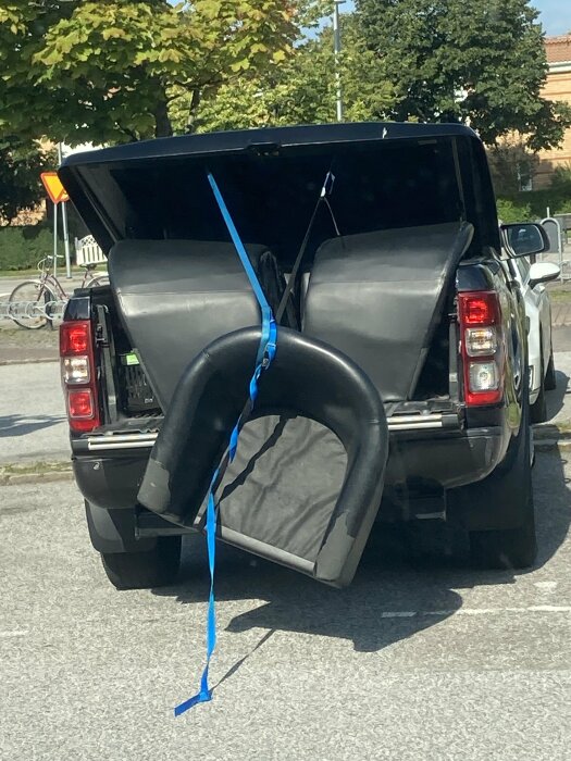 En stor bildäck säkrad med blå band genom en öppen baklucka på en SUV på en parkering.