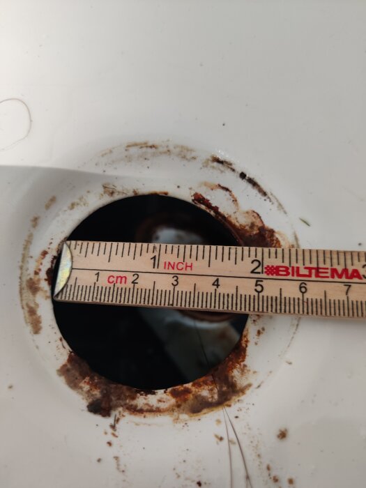 Måttstock över rostig öppning på vit yta mäter diameter; mätaren visar centimeter och tum.