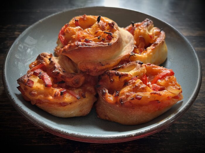 Ostiga pizza-rolls med tomat och röd lök på en grå tallrik mot mörkt träbord.