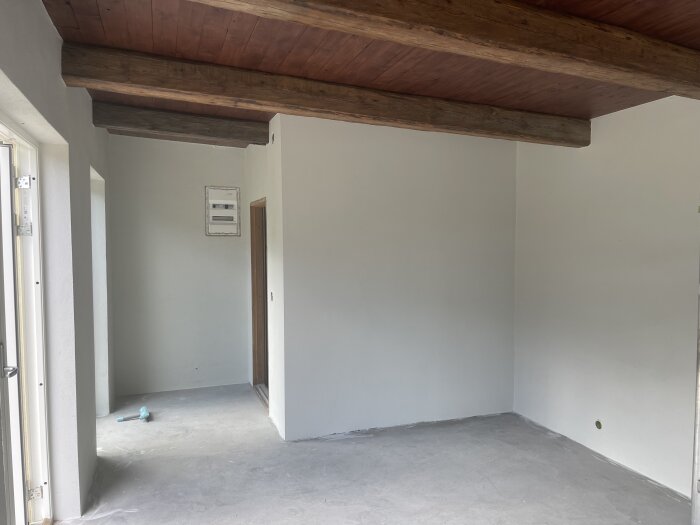Tomt rum, träbjälkar i taket, vitmålade väggar, betonggolv, elskåp på vägg.